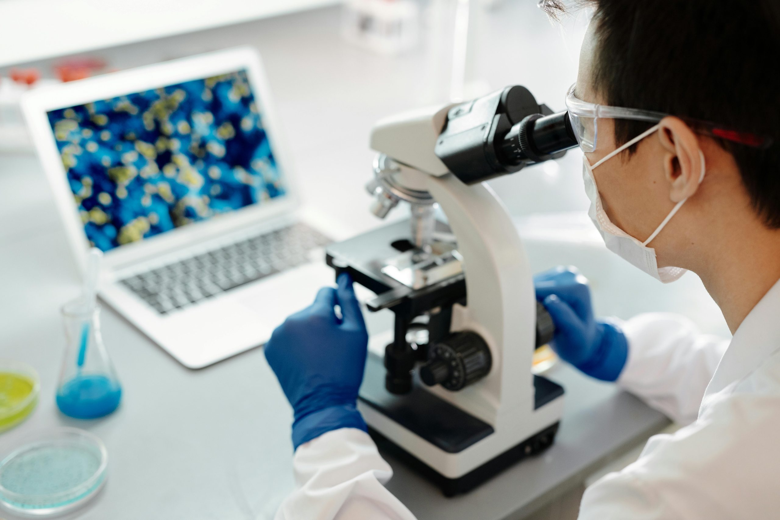 Scientist examining samples under a microscope in a lab with laptop display.